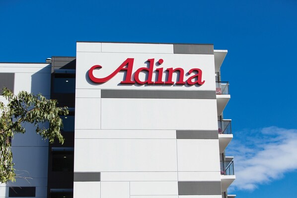 Ein Gebäude mit einem Schild, auf dem in roten Buchstaben „Adina“ steht, vor weißem Hintergrund.