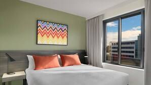 A hotel room with a large bed, a nightstand, a modern lamp, a colorful abstract painting, and a large window with a view of buildings. - Adina Apartment Hotel Sydney Airport (Mascot)