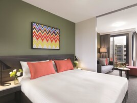 A hotel room with a large bed, a colorful artwork on the wall, a nightstand with a lamp, and a view of the city through the window.