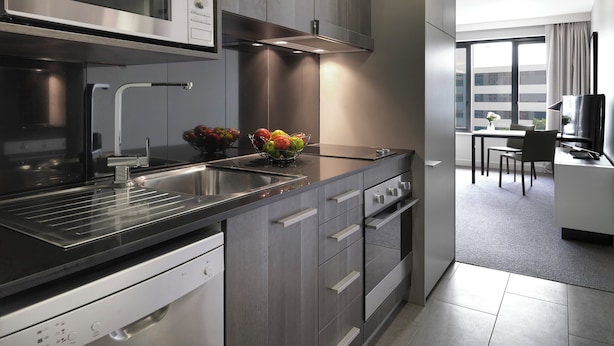 A modern kitchen with a built-in microwave, oven, and sink. There is a fruit bowl on the counter and a dining area with a table and chairs in the background. - Adina Apartment Hotel Sydney Airport (Mascot)