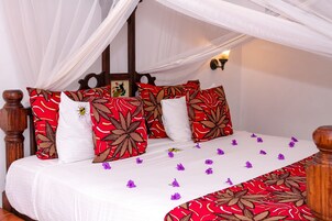 Una cama con dosel, almohadones blancos y rojos con estampado, y una colcha blanca adornada con flores moradas.