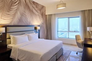 A hotel room with a large bed, a desk with a chair, a large window with a city view, and a wall with a leaf pattern. - Marriott Hotel Production City, Dubai (Dubai)