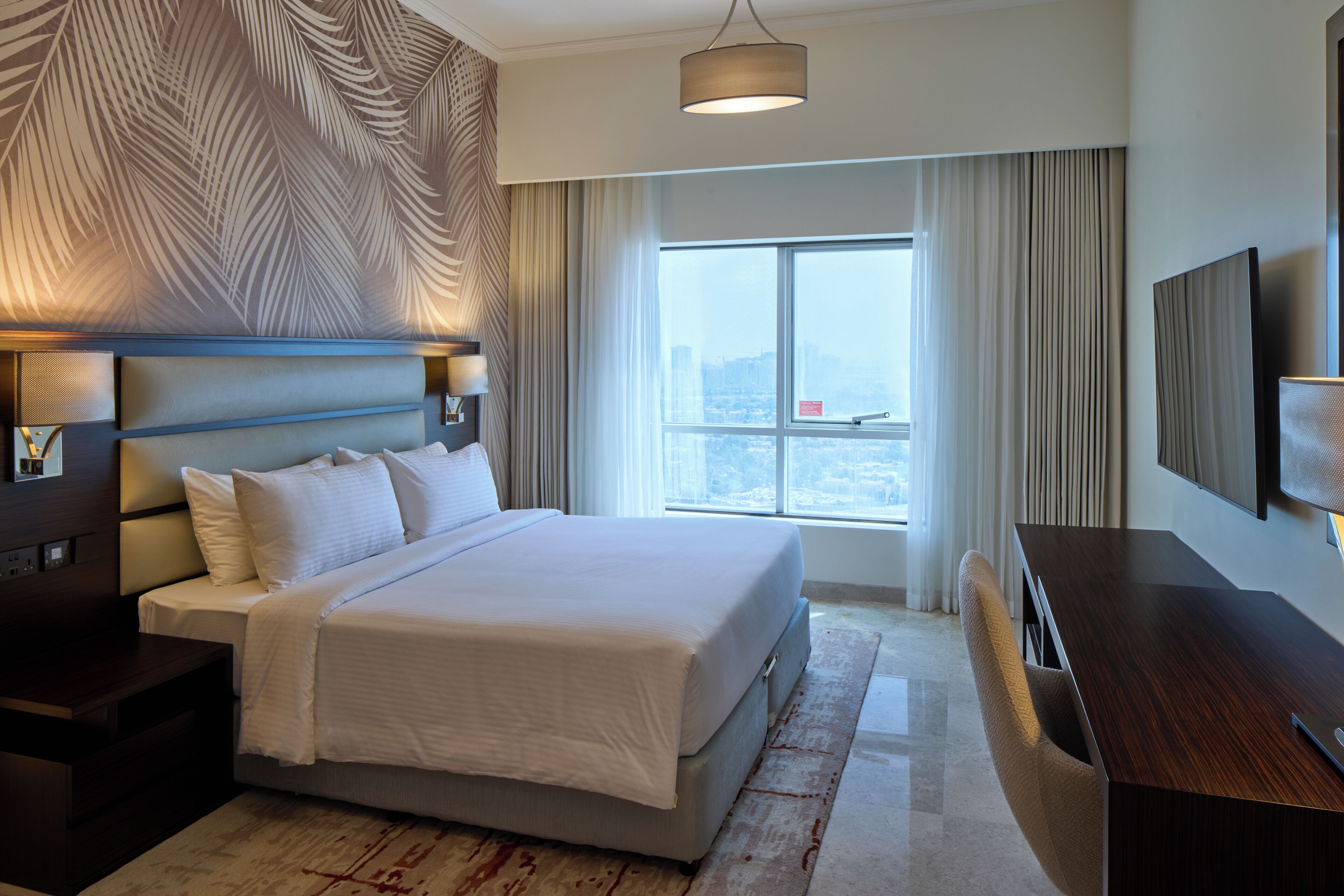 A modern hotel room with a large bed, a desk, a TV, and a city view.