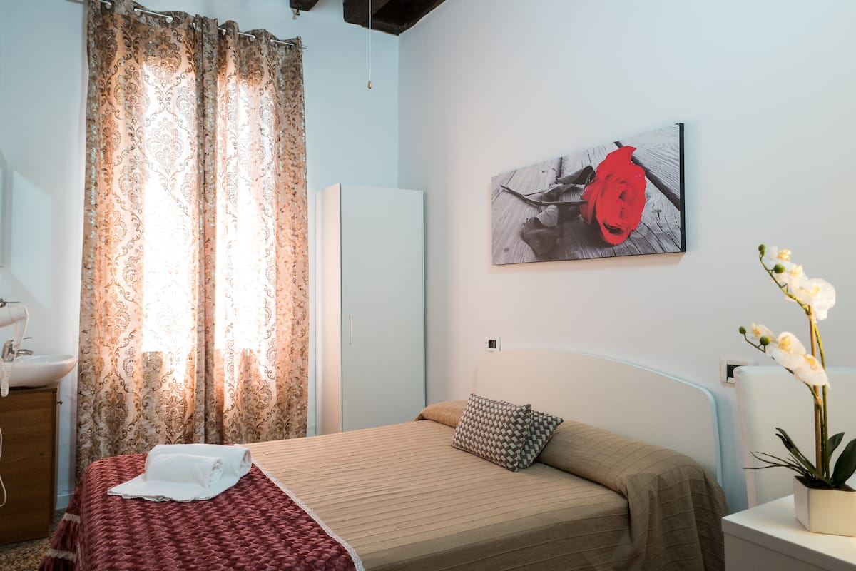 A bedroom with a bed, a bedside table with a lamp, a potted orchid, a painting on the wall, and a window with curtains.