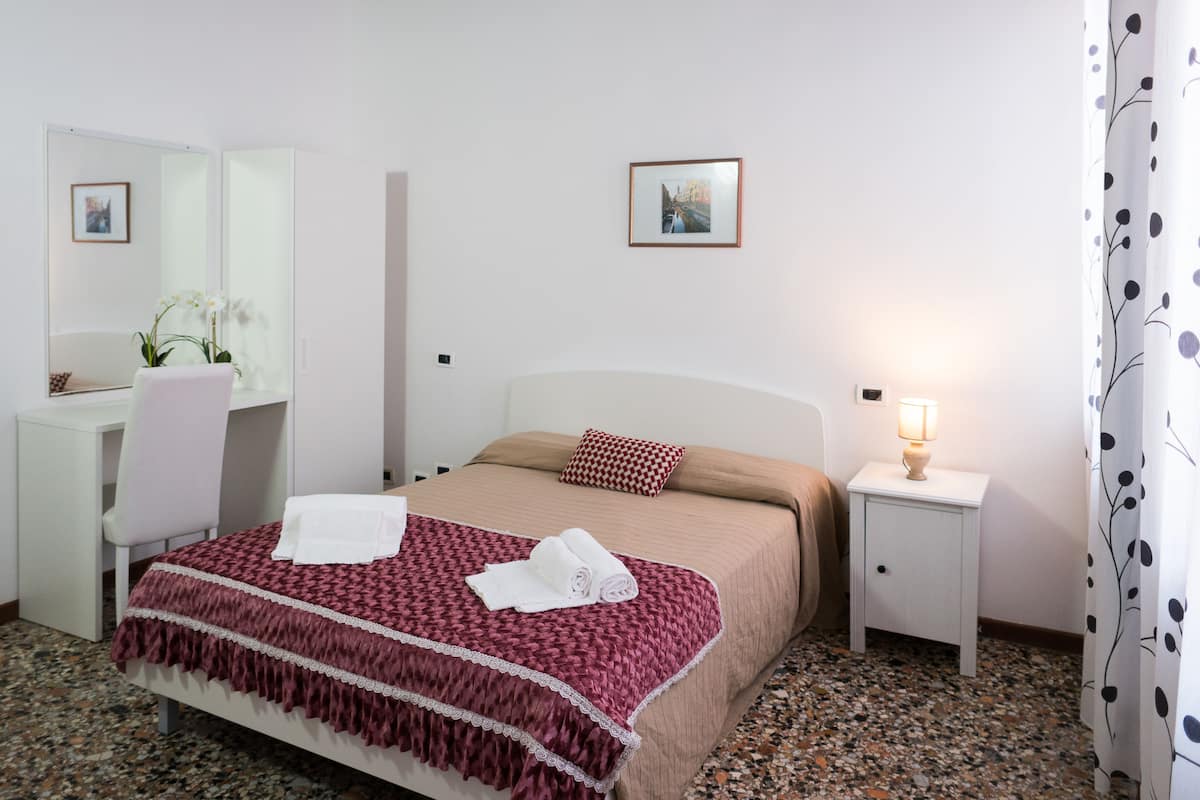 A neatly arranged hotel room with a bed, a desk, a chair, a nightstand with a lamp, and a mirror.