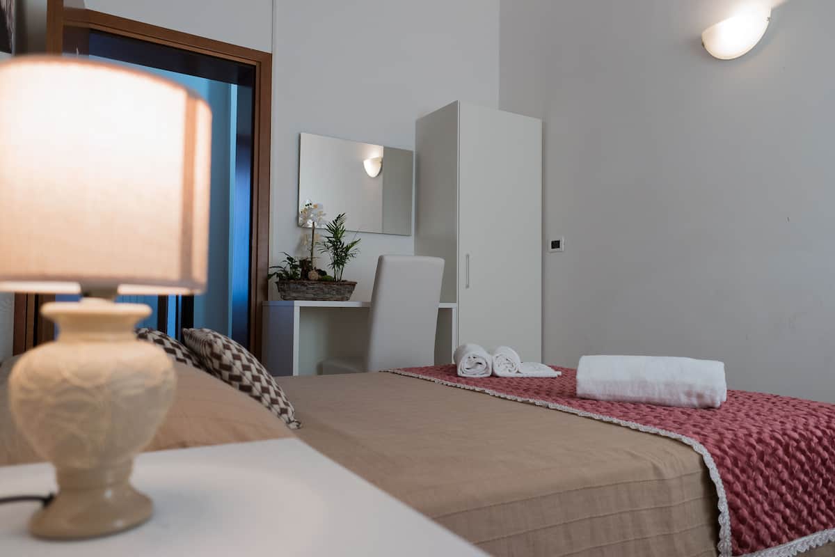 A hotel room with a bed, a bedside table with a lamp, a mirror, a wardrobe, and a red blanket.