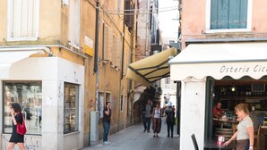 A narrow street with buildings on either side, a café with outdoor seating, and pedestrians walking by. - Backpackers House Venice - Hostel (Venice)