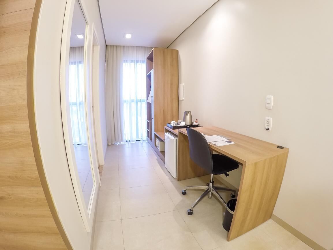 Une chambre d’hôtel moderne avec un bureau en bois, un fauteuil de bureau noir, une petite kitchenette et une vue sur un couloir avec une fenêtre.