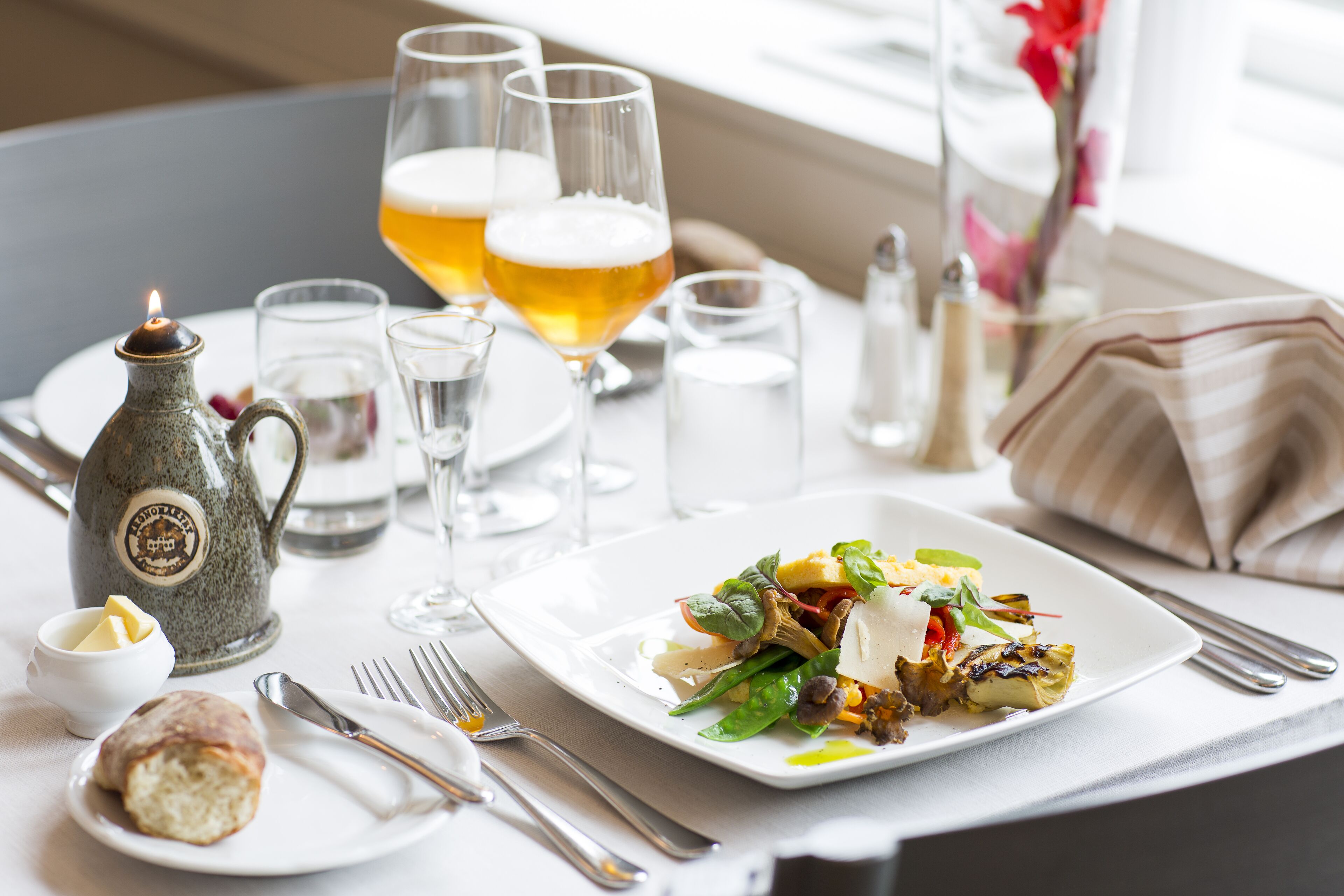 A dining table with a plate of food, glasses of beer, a teapot, and cutlery.