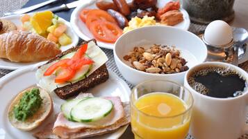 Esmorzar amb croissants, tomàquets a rodanxes, cogombre, pernil, un bol de granola i un got de suc d'aranja.