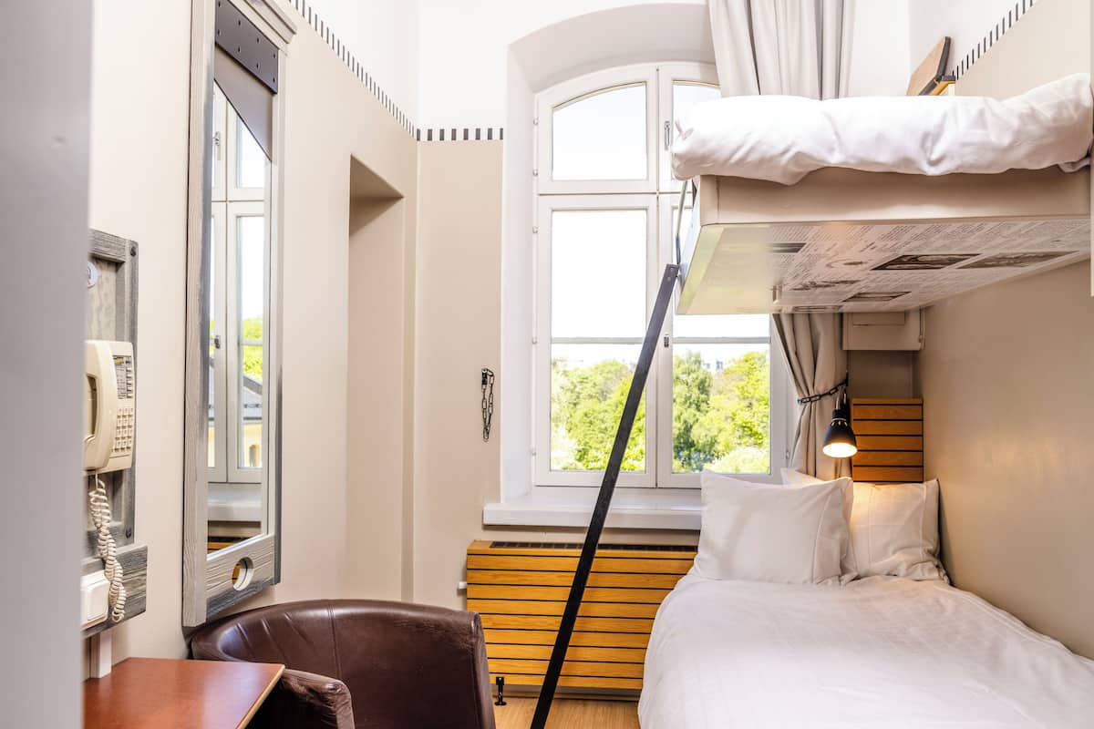 A compact hotel room with a bunk bed, a wooden desk, a telephone, and a window with curtains.