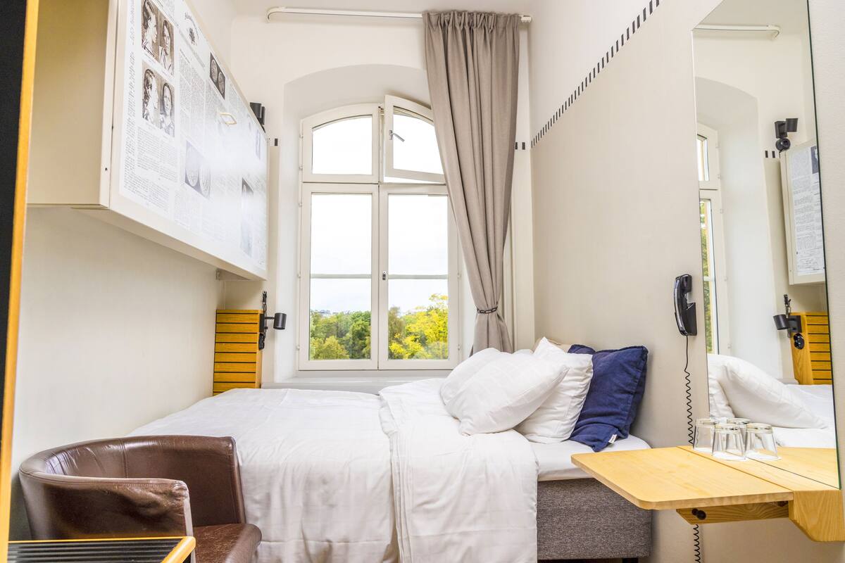 A bedroom with a bed, a desk, a chair, a window with curtains, a phone, and a wall-mounted mirror.