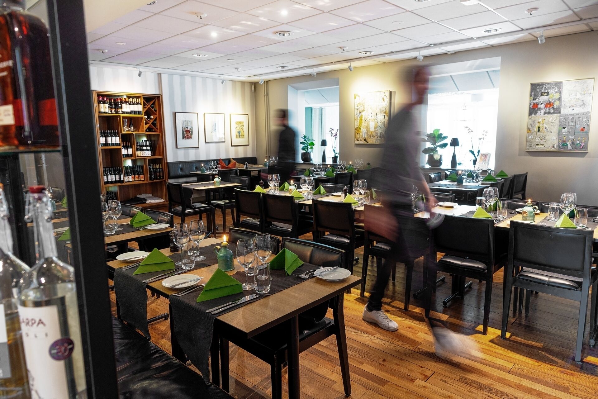 A restaurant with multiple tables set for dining, a wine rack in the background, and a person walking by.