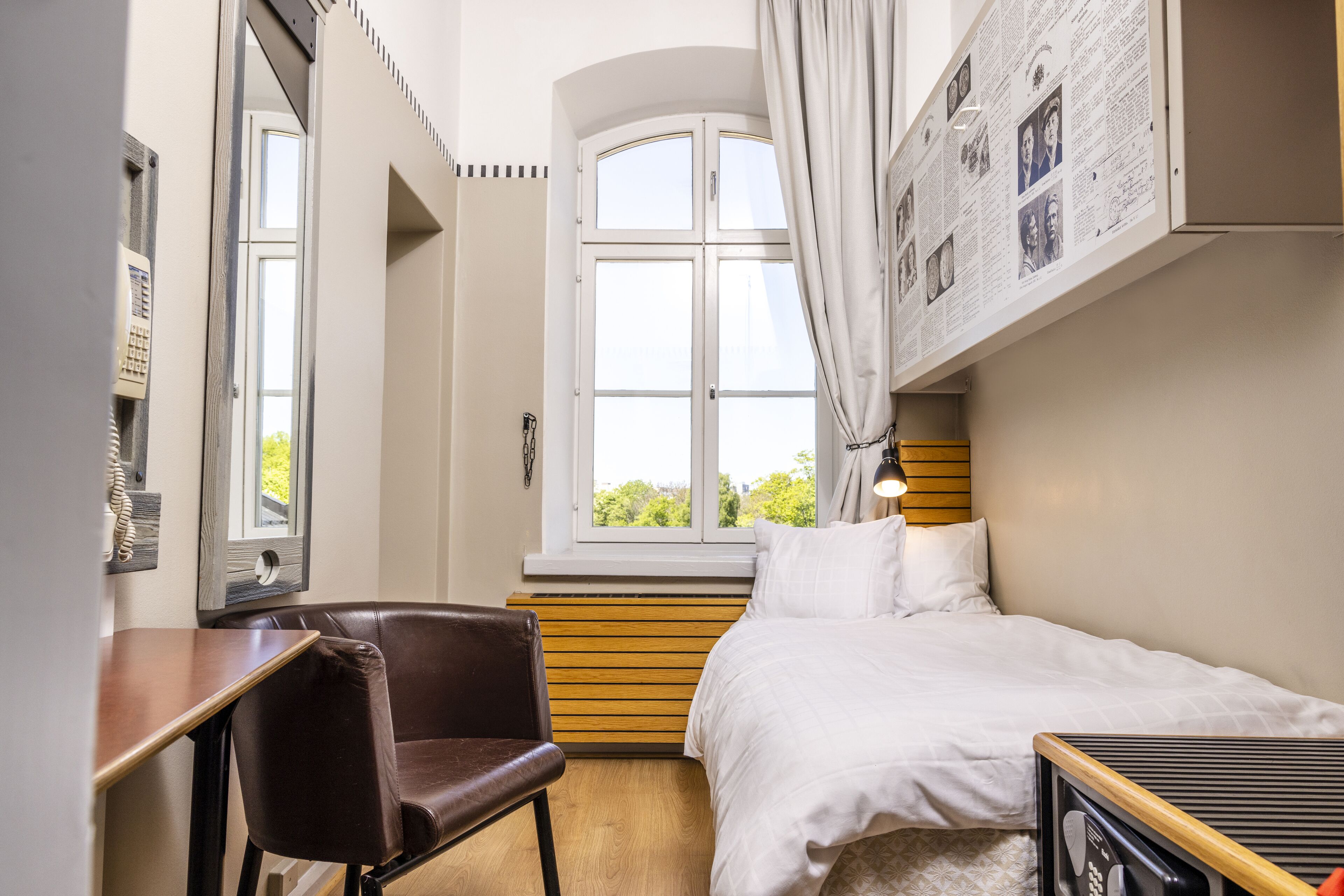 A hotel room with a bed, a desk, a chair, a window, and a wall with photos.