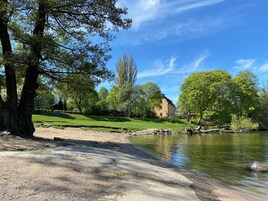 Escena junto al río con una casa, árboles y cielo despejado.