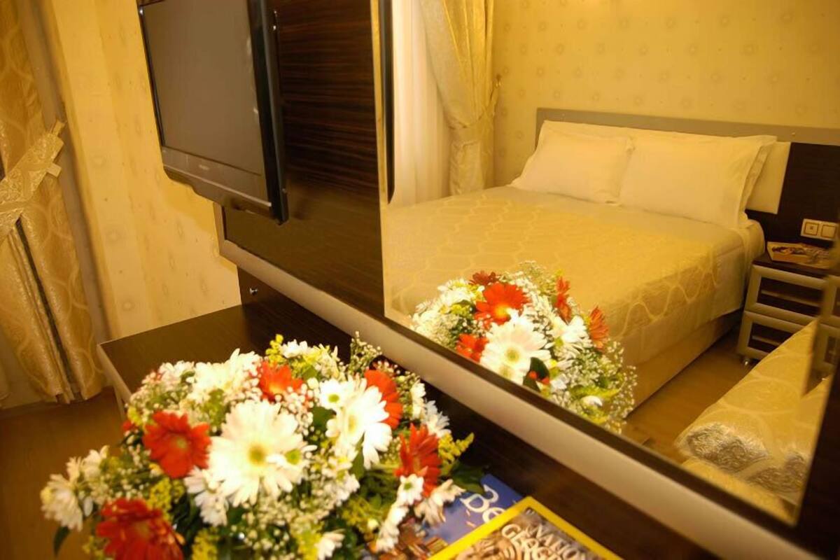 A hotel room with a bed, a TV, a bedside table with a floral arrangement, and a mirror reflecting the room.