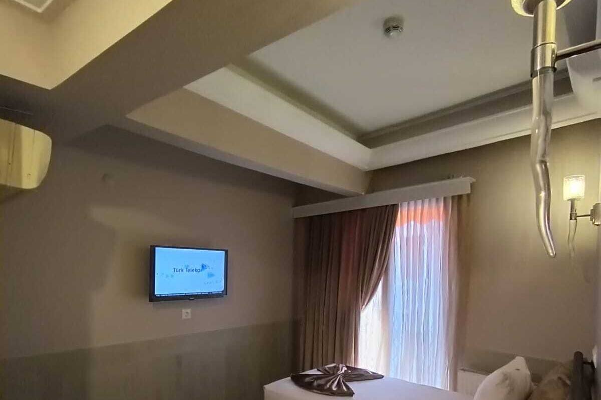 A hotel room with two beds, a flat-screen TV mounted on the wall, a window with curtains, and a wall-mounted lamp.