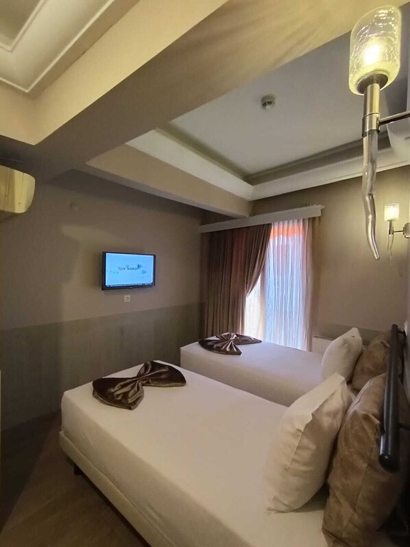 A hotel room with two beds, a flat-screen TV mounted on the wall, a window with curtains, and a wall-mounted lamp.