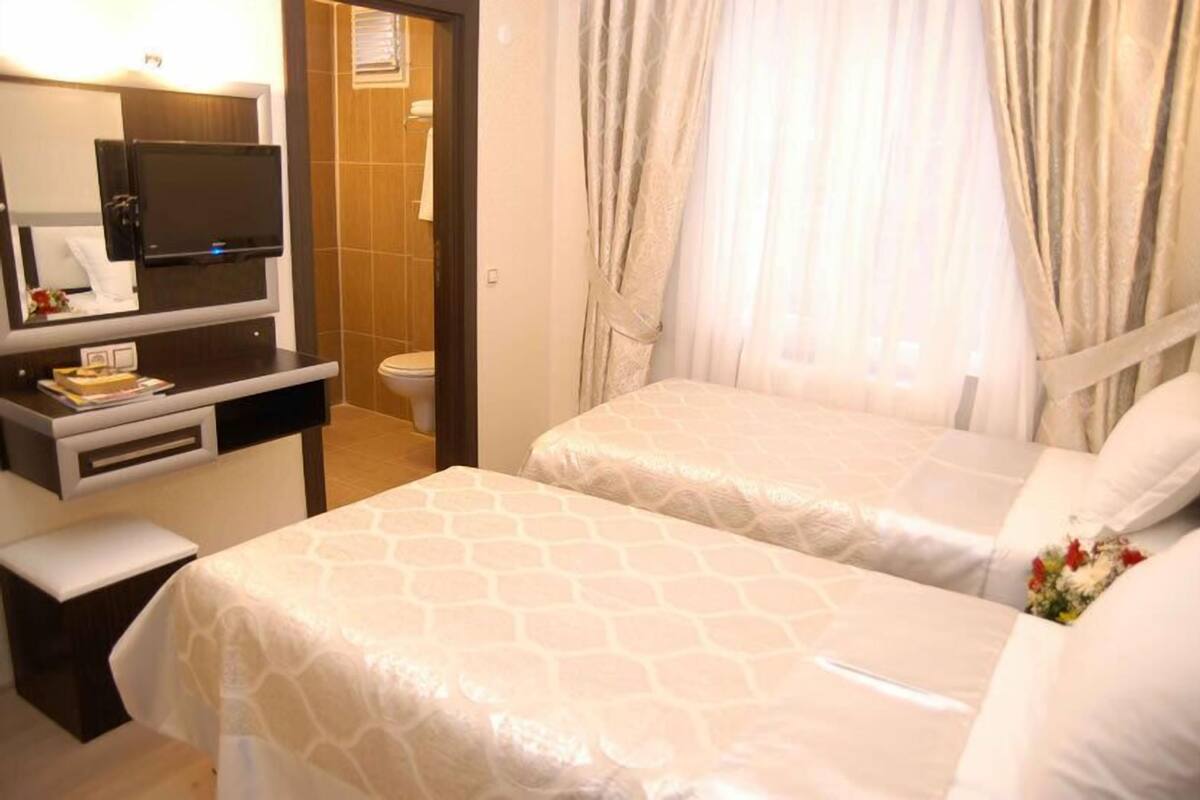 A hotel room with two beds, a TV mounted on the wall, a small desk with a chair, and a shower area visible through a glass door.