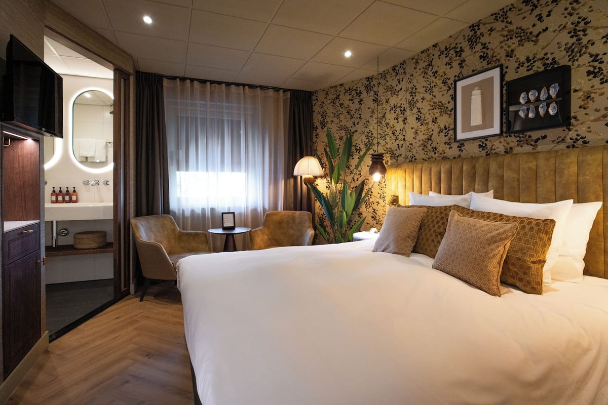 A hotel room with a large bed, a desk with a chair, a mirror, and a wall-mounted shelf with decorative items.