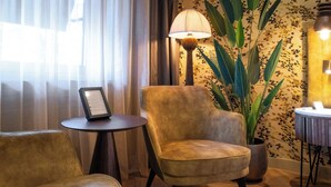 Een kamer met een houten vloer, een bank, een stoel, een klein tafeltje met een lamp en een plant in pot.