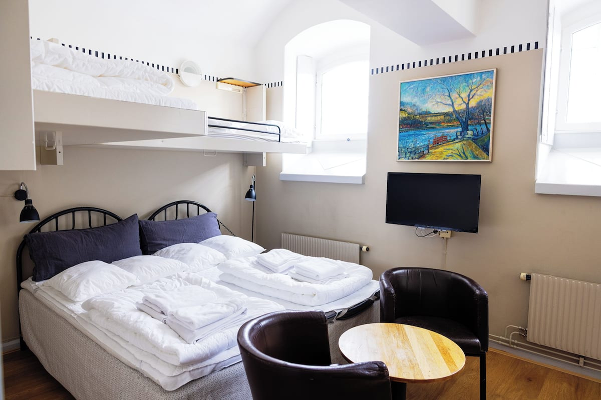 A hotel room with a bunk bed, a single bed, a small round table, a black leather chair, a painting on the wall, and a television.