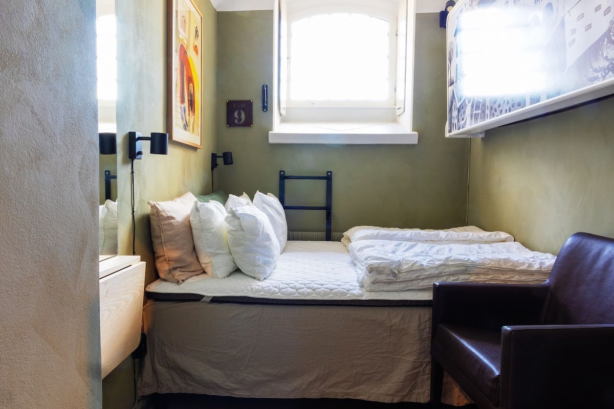A small, single-bed room with a window, a wall-mounted light, and a chair.