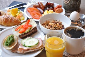 Une table dressée pour le petit-déjeuner avec des croissants, des tomates en tranches, des concombres, du jambon, un bol de granola et un verre de jus d’orange.