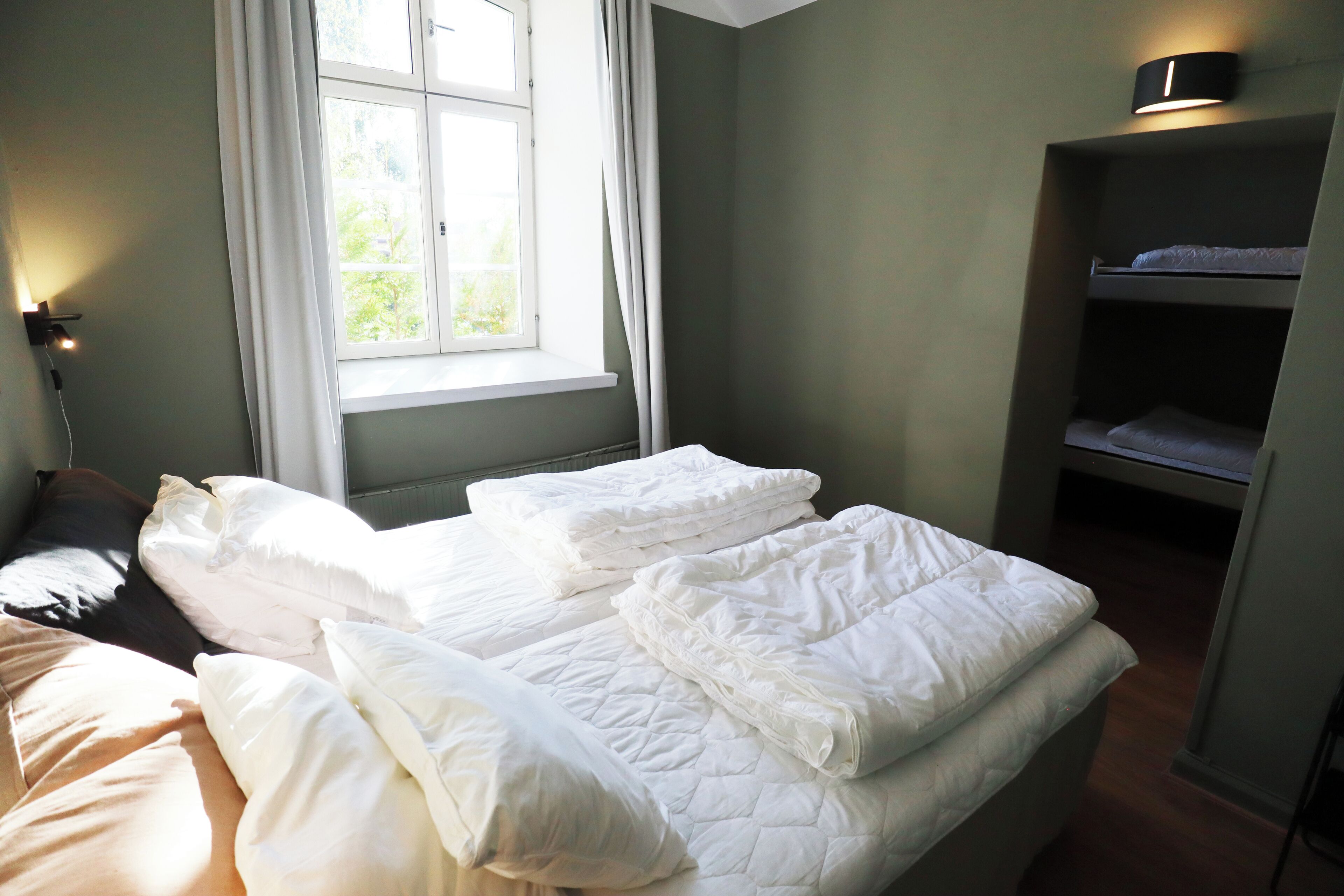Una habitación con dos camas, una ventana con cortinas, una lámpara de noche y una repisa con lámpara.