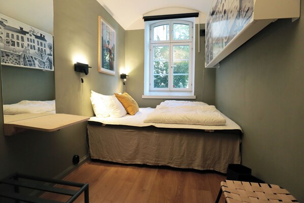 A bedroom with a bed, a window, a wall-mounted shelf, and a bedside table. - STF Långholmen Hostel (Stockholm)