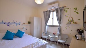 Un dormitorio con cama, armario, una pequeña mesa con sillas y una ventana con cortinas.