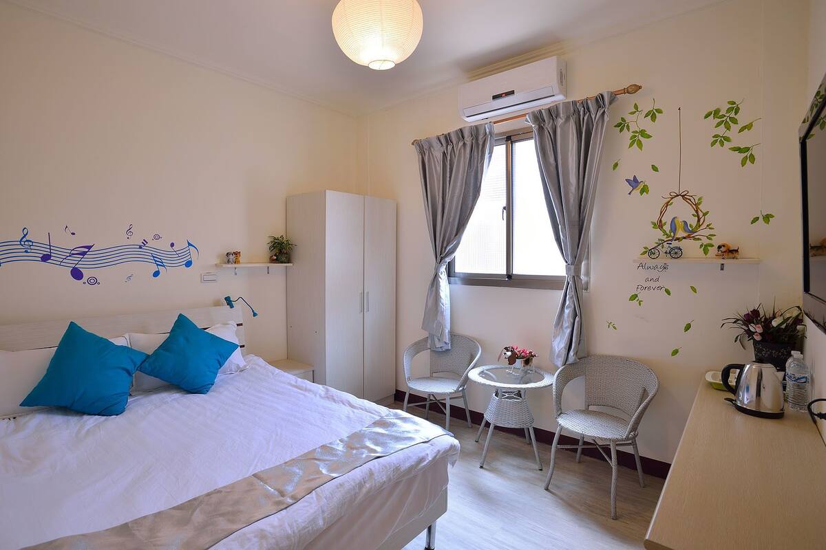 A bedroom with a bed, a wardrobe, a small table with chairs, and a window with curtains.