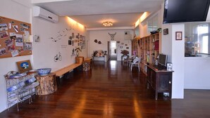Un pasillo espacioso con piso de madera, un tablón de anuncios con varias fotos y notas, una estantería llena de libros y un televisor montado en la pared.