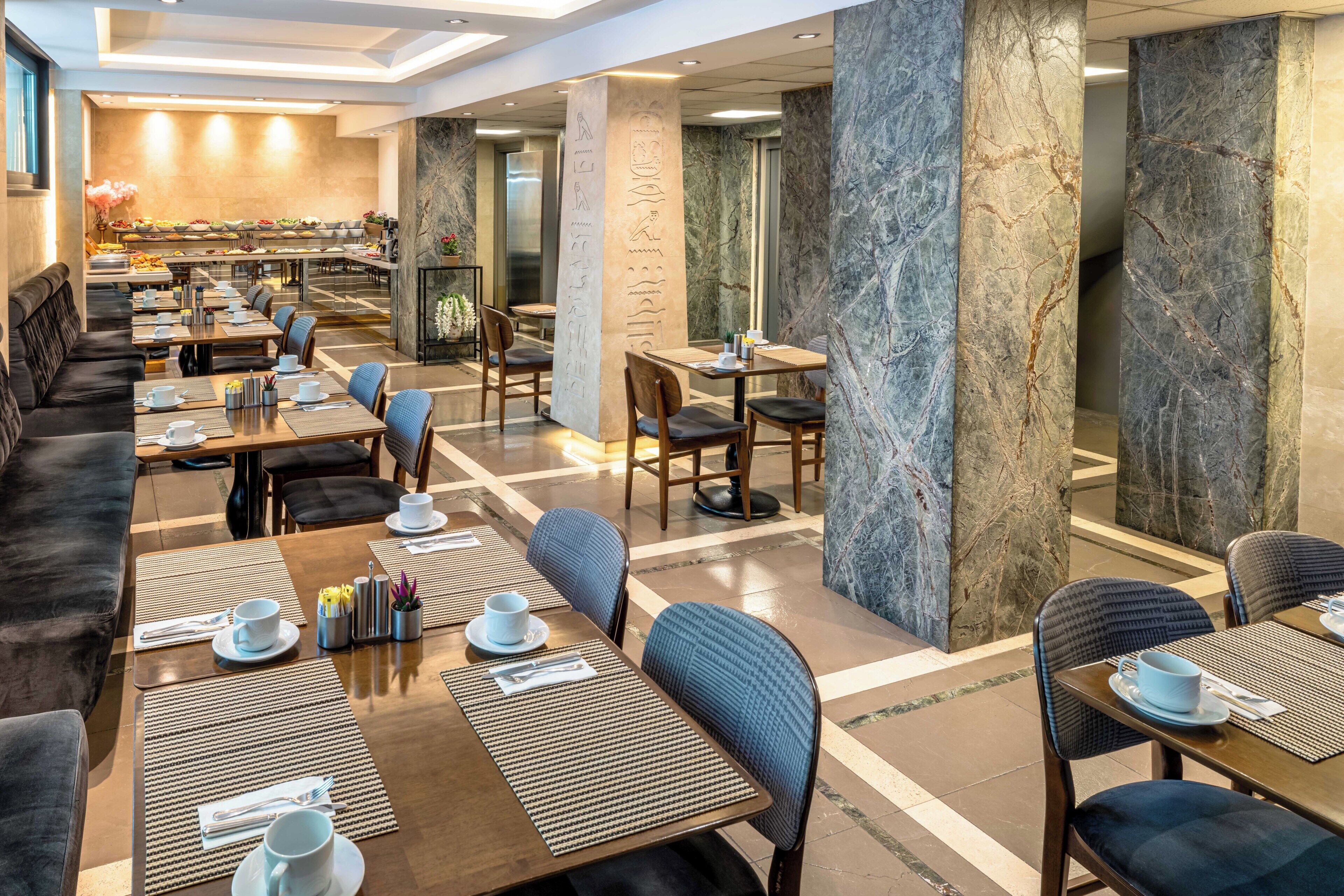 Ein Hotelrestaurant mit mehreren Tischen und Stühlen, einem Buffet im Hintergrund und Marmorsäulen.