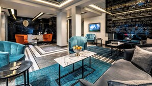 En moderne hotellobby med en receptionsdisk i marmor, siddepladser i teal og grå farver, et stort vægmonteret TV og et mønstret vægdesign.