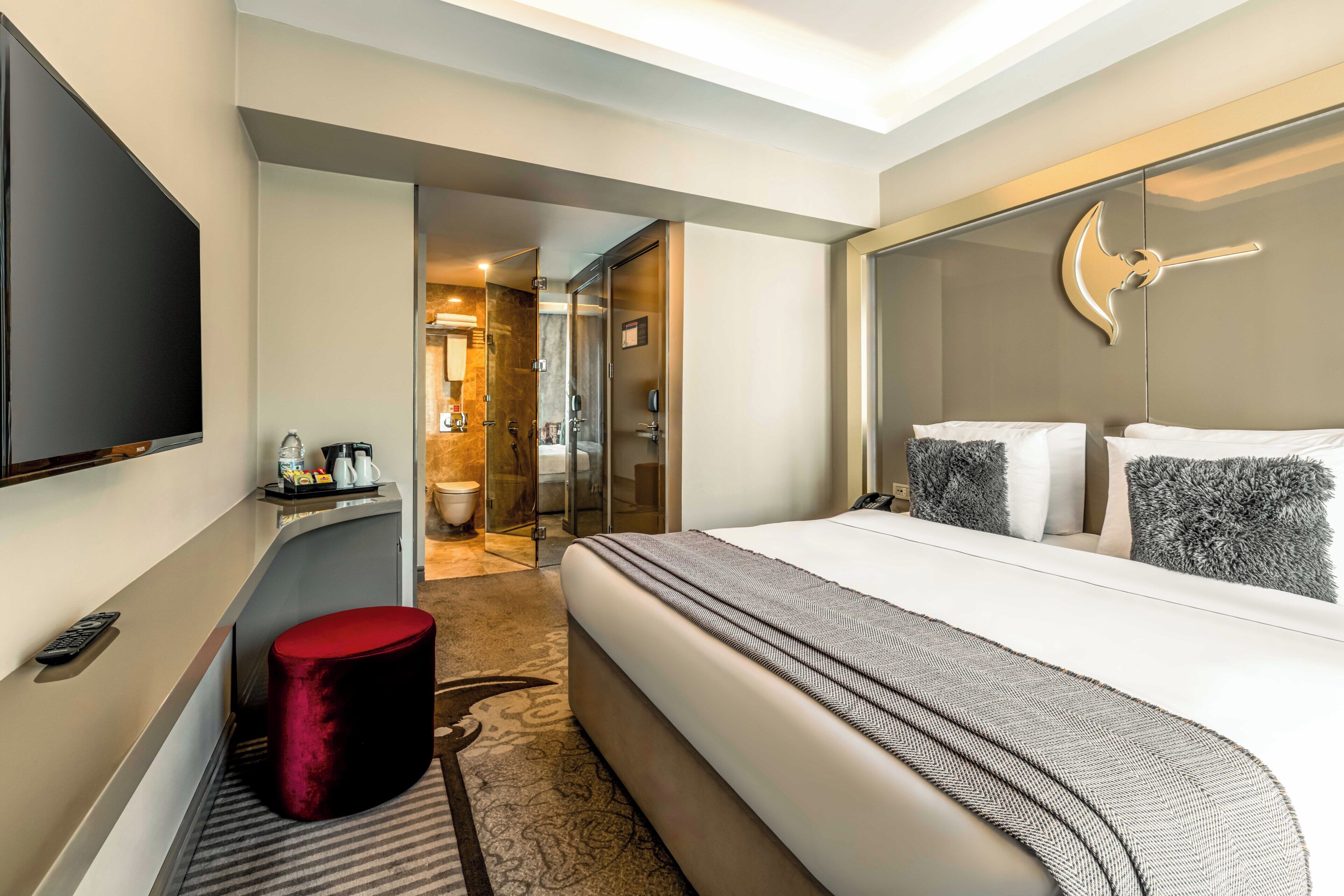 A hotel room with a large bed, a TV, a red ottoman, and a mirror with a decorative wall piece.