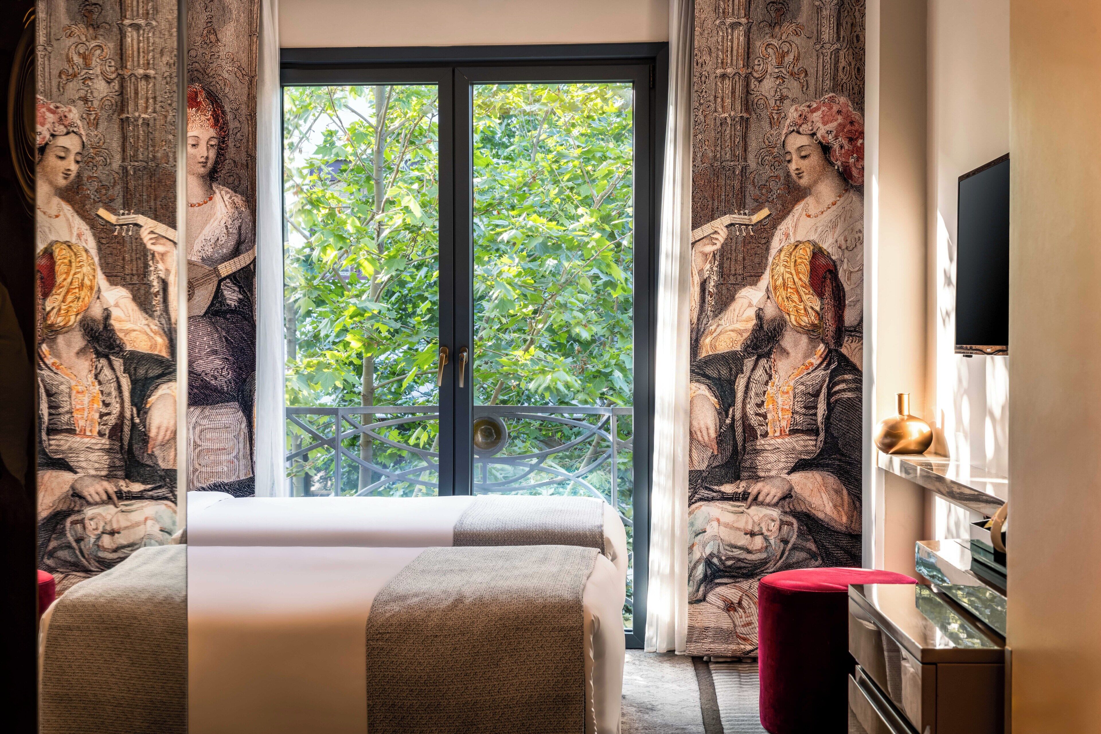 Een hotelkamer met een groot bed, gordijnen met een klassiek schilderij, een balkon met uitzicht op bomen en een flatscreen televisie op het dressoir.