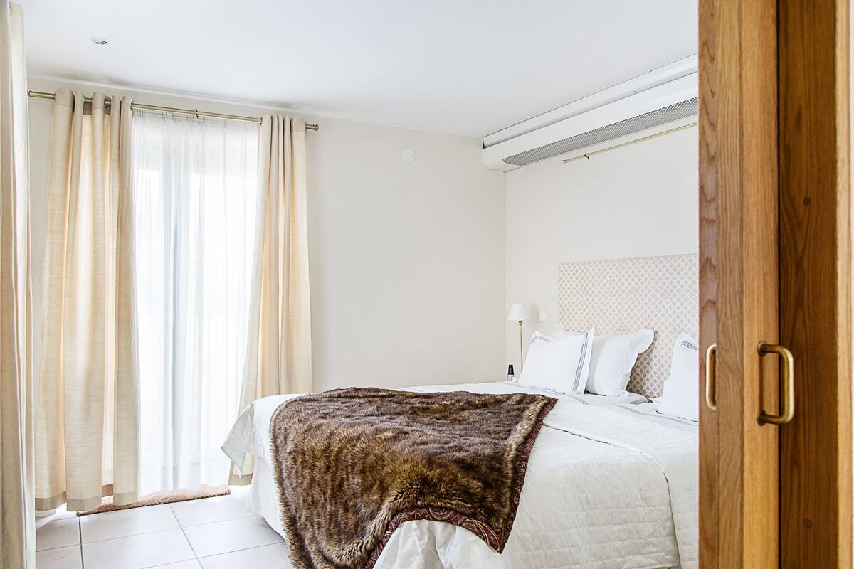 Quarto de hotel com cama, cortinas, mesa de cabeceira com abajur e porta de madeira.