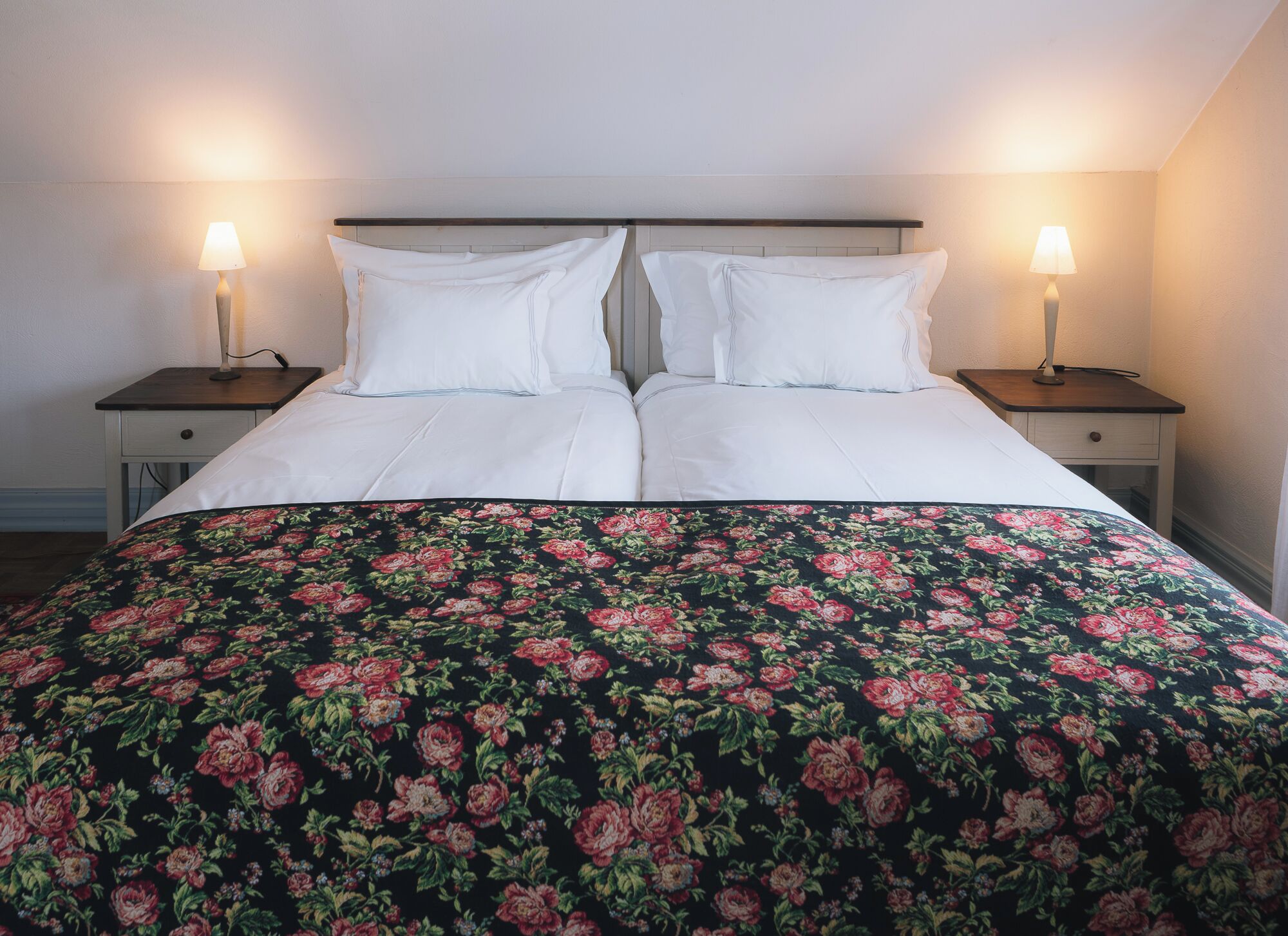 A double bed with white linens and a floral-patterned bedspread. There are two bedside tables with lamps on either side of the bed.