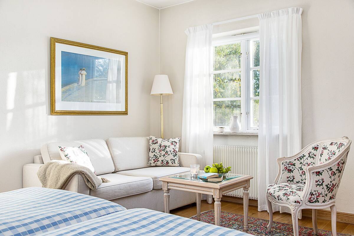Um quarto com sofá, poltrona, mesa com um livro e maçãs, abajur e um quadro emoldurado.