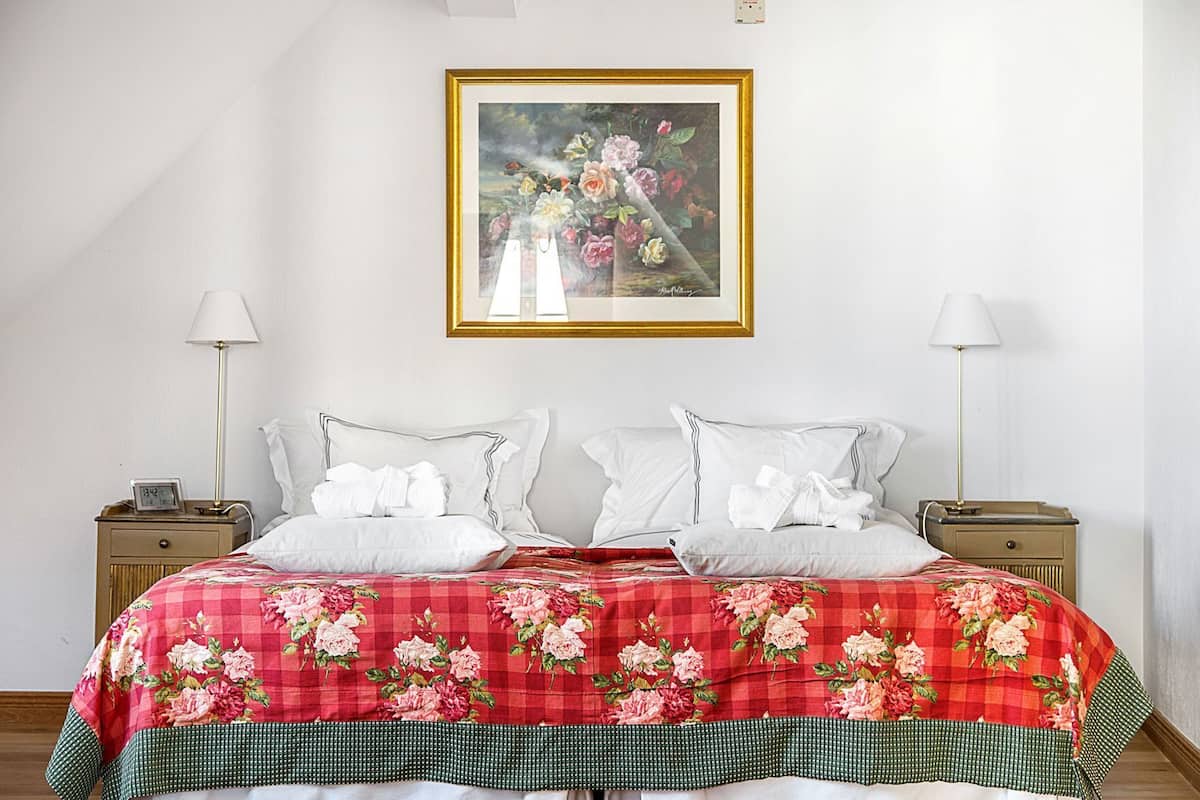 Uma cama bem arrumada com colcha floral, duas mesas de cabeceira com luminárias, um quadro acima da cama e um relógio em uma das mesas.