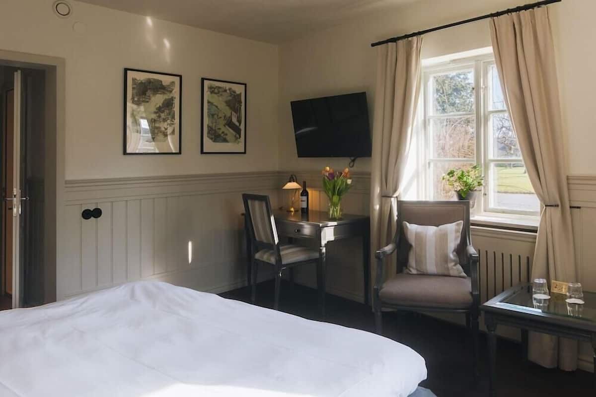 Quarto de hotel com cama, escrivaninha com cadeira, televisão e duas fotos emolduradas na parede.
