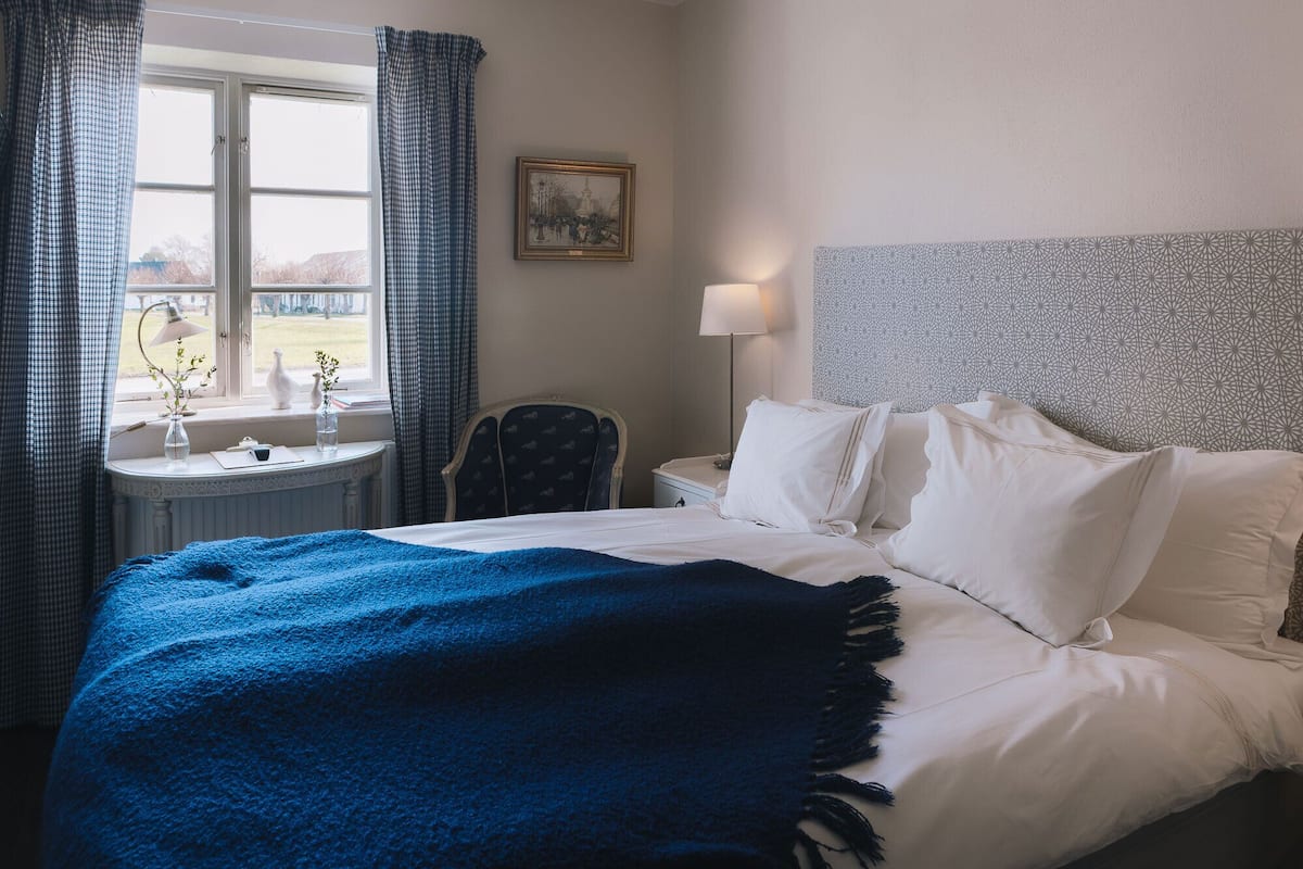 Quarto de hotel com cama, mesa de cabeceira, luminária, cadeira, janela com cortinas e um quadro na parede.