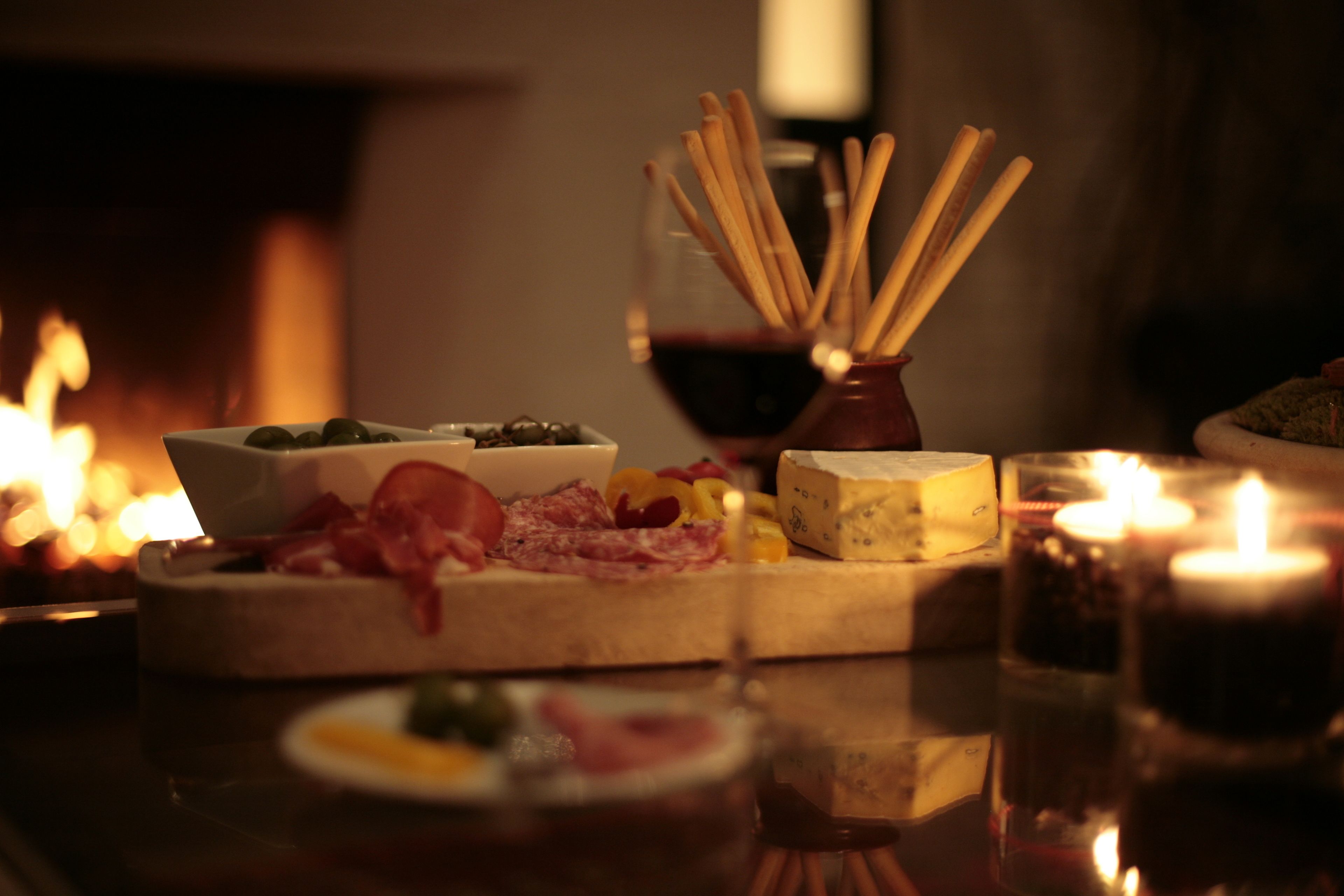 Uma mesa com variedade de queijos, carnes e azeitonas, acompanhada por velas e um copo de vinho tinto.