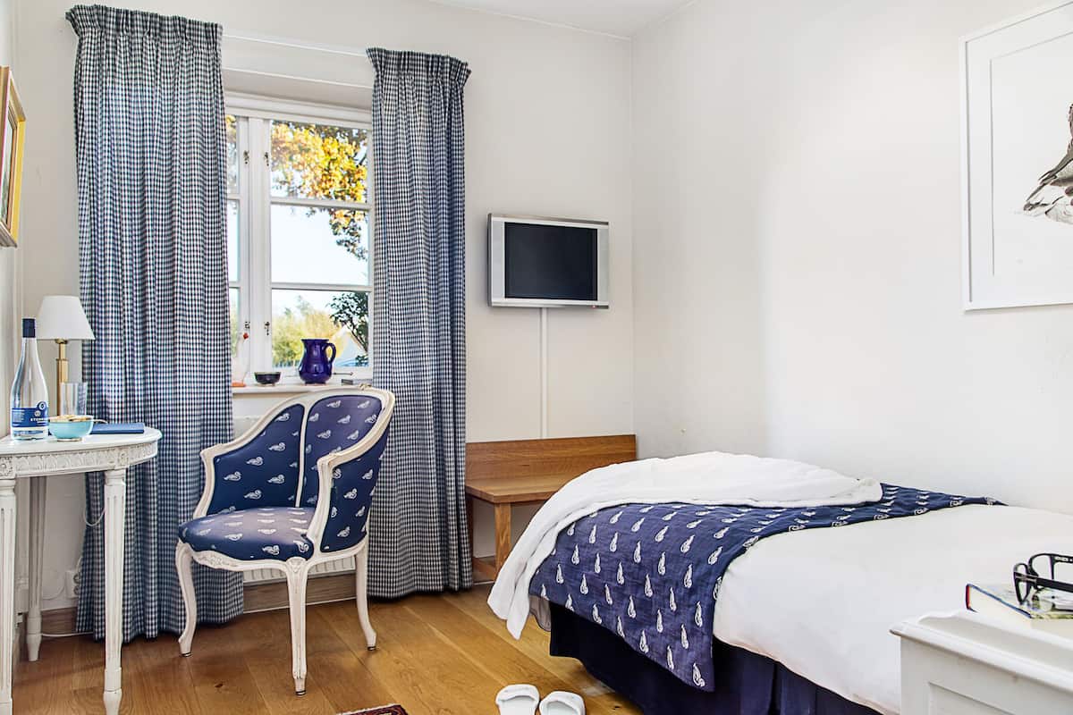 Quarto com cama, escrivaninha, cadeira, televisão e janela com cortinas.