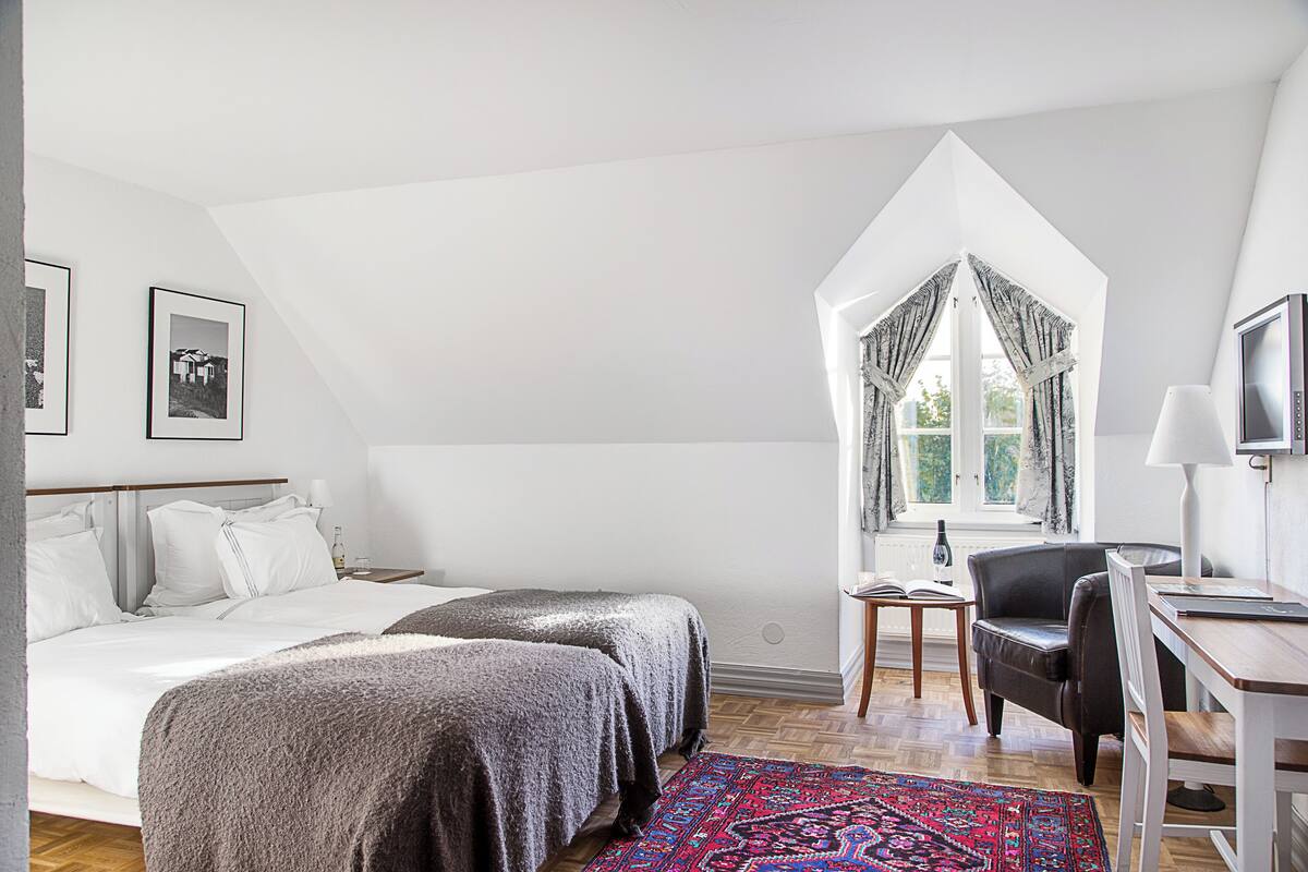 Quarto com duas camas, uma escrivaninha, uma cadeira, janela com cortinas e um tapete estampado.