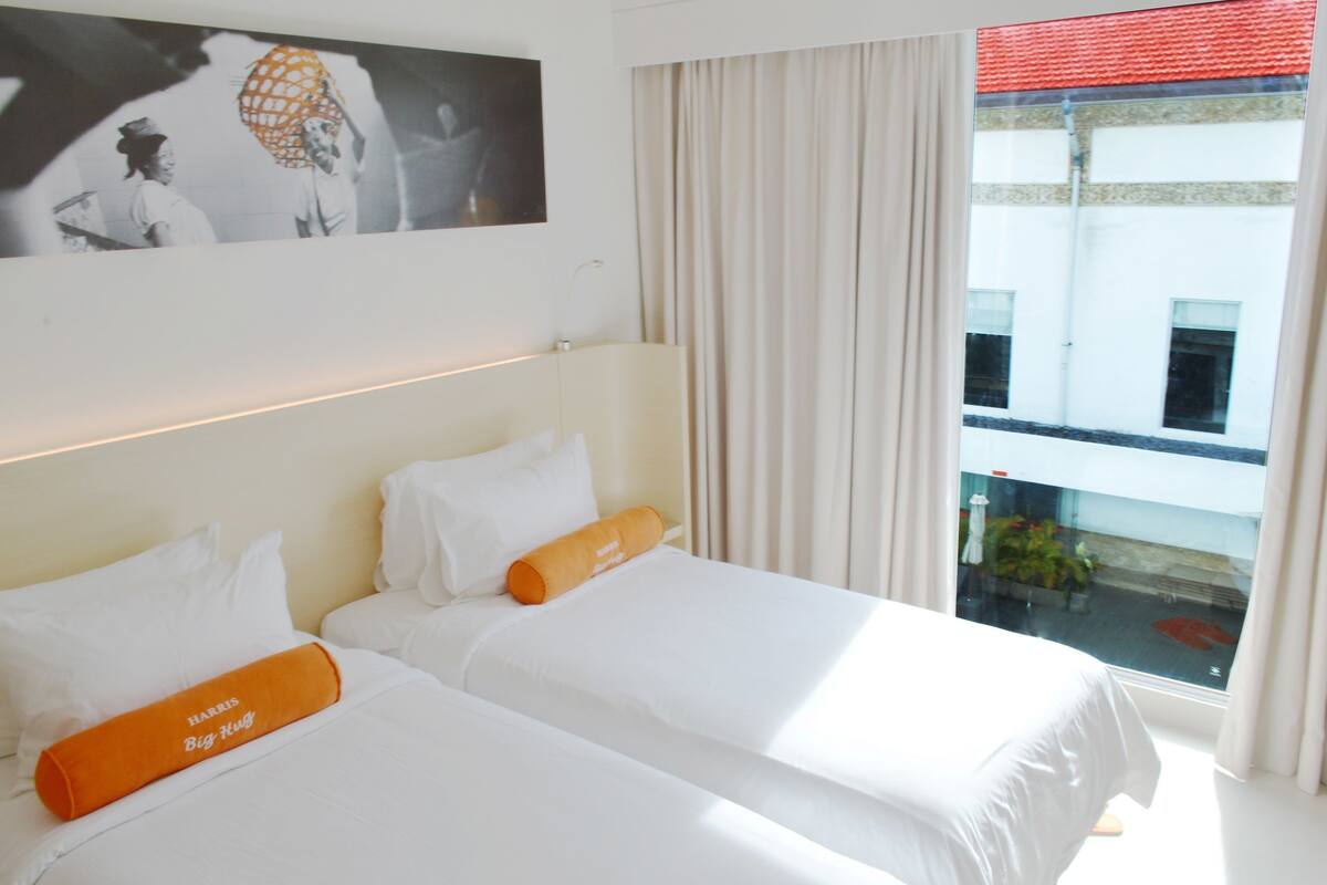 A hotel room with two beds, white bedding, a large window with curtains, and a view of a building outside.