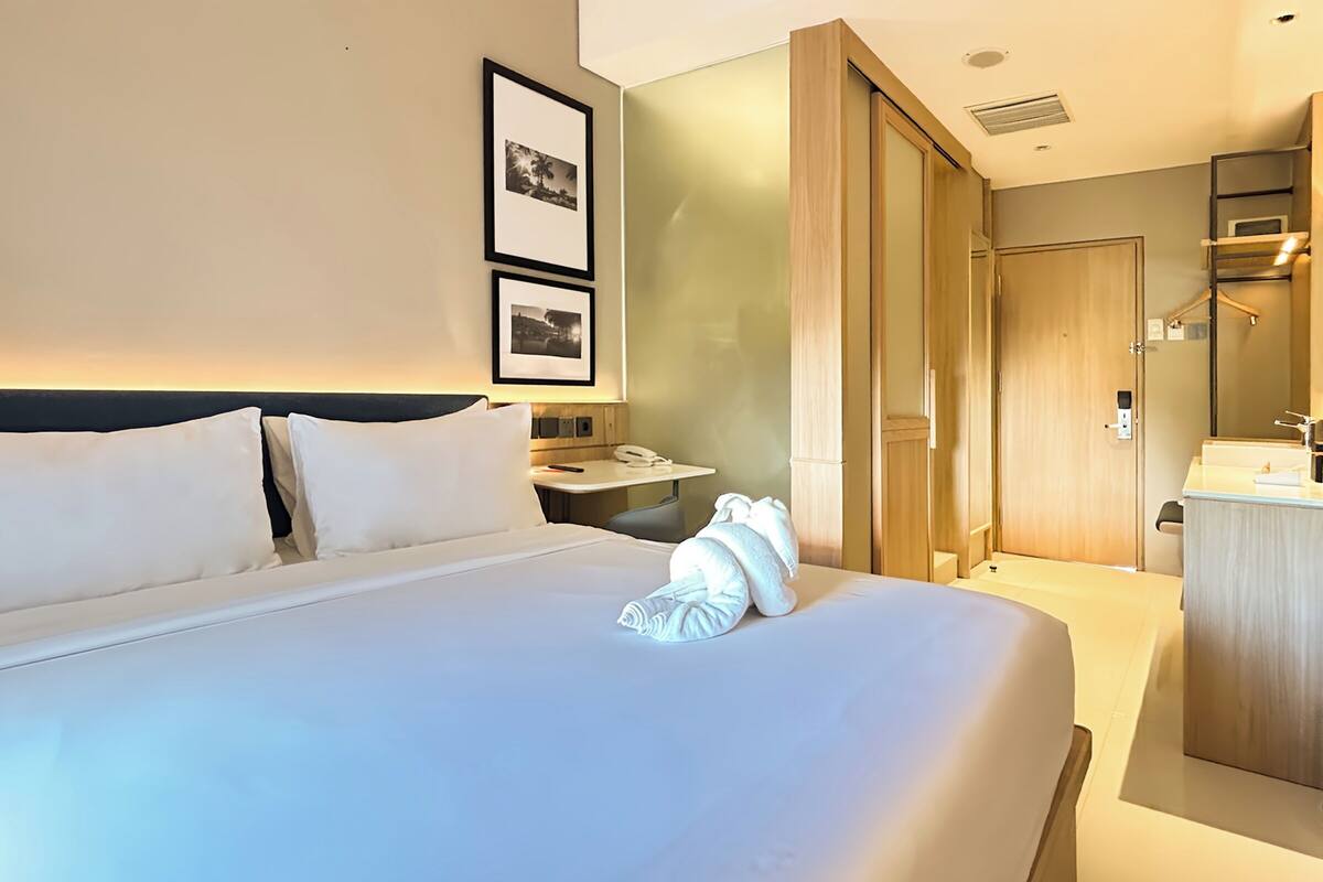 A hotel room with a neatly made bed, a bedside table with a phone, and a bathroom with a sink and mirror.