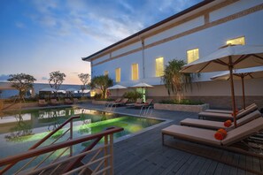 A pool area with lounge chairs and umbrellas, a building with large windows, and a clear sky. - HARRIS Hotel & Conventions Denpasar Bali (Denpasar)