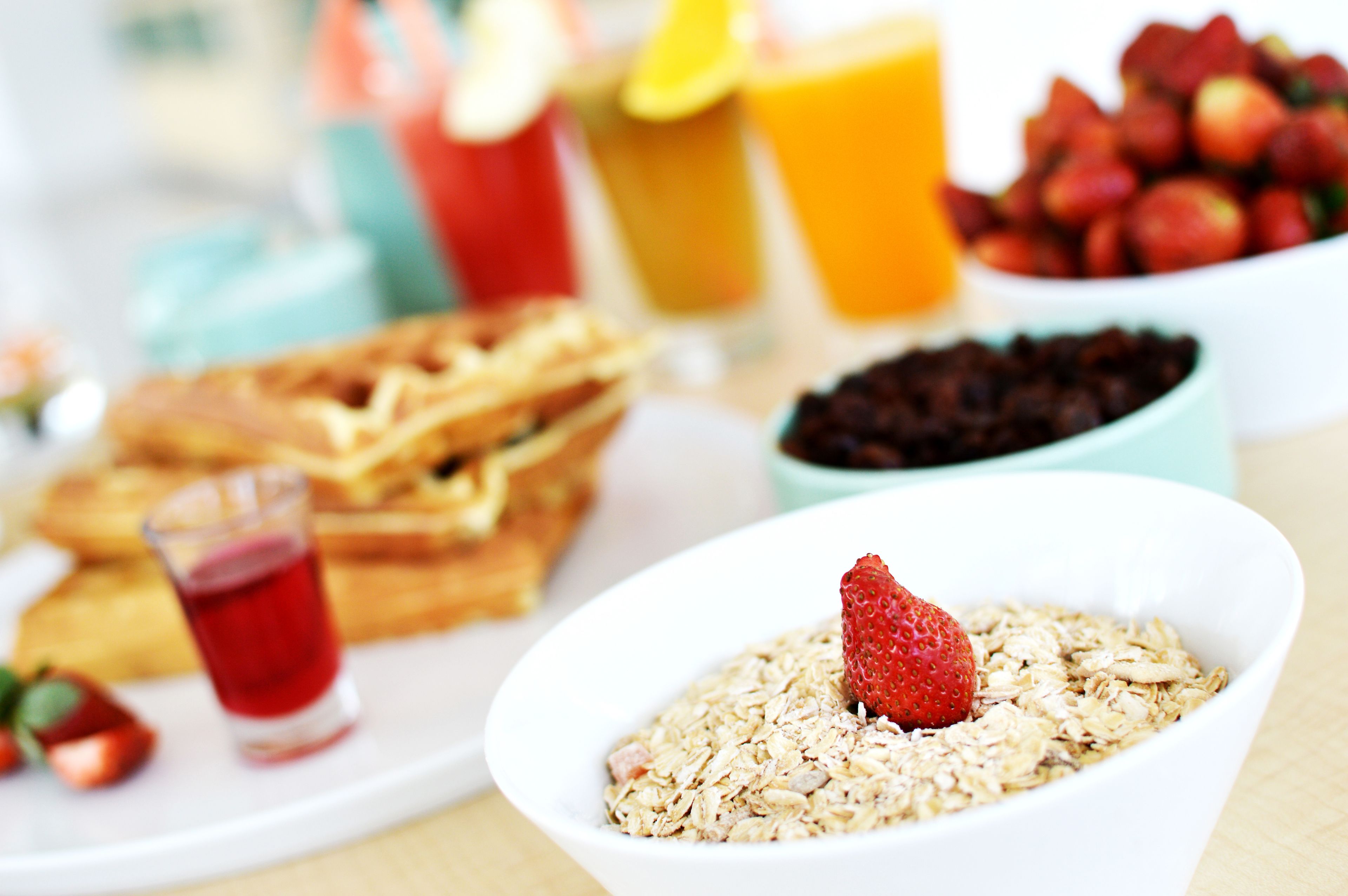 Une table mise pour le petit-déjeuner avec des gaufres, un bol de porridge agrémenté d’une fraise, un verre de jus rouge et un bol de fraises.